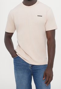 Hombre con camiseta beige con el logo "BARROW" en el pecho y pantalones vaqueros azules, de pie con una mano en el bolsillo contra un fondo liso.