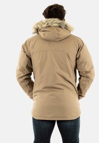 Giacca isolante beige con cappuccio foderato in pelliccia, presenta una texture liscia e un orlo leggermente curvo, indossata sopra jeans di denim scuro.