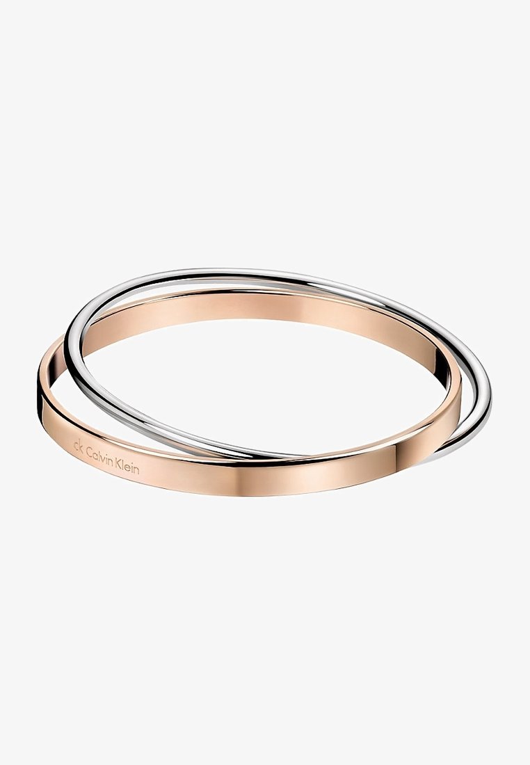 Deux bracelets fins entrelacés, l’un en or rose avec gravure Calvin Klein et l’autre en argent brillant, sur fond blanc.