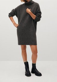 Abito maglione oversize grigio scuro con colletto alto e polsini a coste, abbinato a stivali neri alla caviglia e calzini crema.