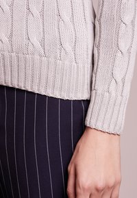 Pull gris clair en maille torsadée avec ourlet et poignets côtelés, détail de motif texturé. Assorti à un pantalon rayé bleu marine.