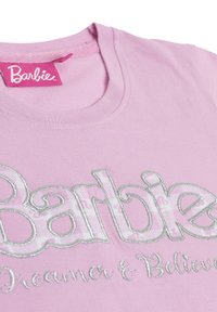 Tela rosa con texto "Barbie" contornado en plateado y bordado más pequeño "Dreamer & Believer" debajo, con una etiqueta rosa de Barbie cerca del cuello.