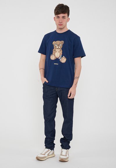 T-shirt blu navy con una grafica di un orso sulla parte frontale. Abbinato a jeans scuri e sneakers beige e bianche. Design semplice, vestibilità casual.