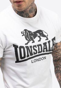 Fehér pamut póló középen fekete oroszlán grafikával és "LONSDALE LONDON" felirattal. Slim fit, rövid ujjú, sima tapintású.