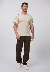 Cremfarbener T-Shirt mit kurzen Ärmeln und schwarzem Seiteneinsatz, kombiniert mit dunkelolivgrünen Hosen. Das Model trägt weiße Sneaker. Glatte Stofftextur.