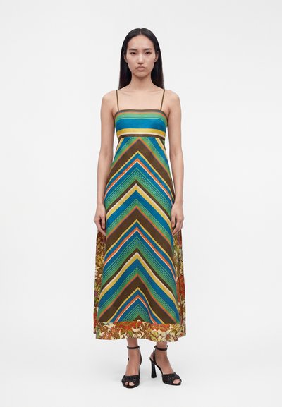 Femme aux longs cheveux foncés portant une robe longue sans manches à rayures multicolores et à motifs floraux avec de fines bretelles et des sandales noires à talons hauts.
