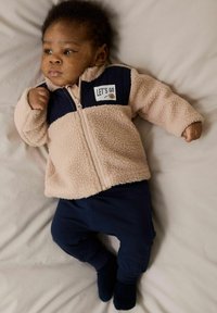 Fleece jack in lichtbeige met marineblauwe accenten, voorzien van een ritssluiting aan de voorkant en een "LET'S GO" graphic. Marineblauwe broek en donkerblauwe sokken maken de outfit compleet.