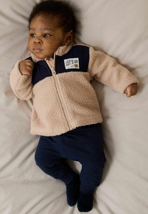 Gaišs beidžs mīkstā jakā ar tumši zilu akcentu, ar priekšējo rāvējslēdzēju un "LET'S GO" grafiku. Tumši zilas bikses un tumši zilas zeķes noslēdz kopējo tērpu.