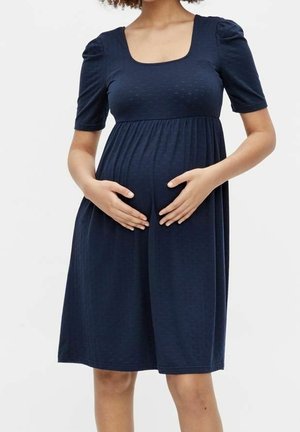 Schwangere Frau trägt ein knielanges, marineblaues Kleid mit kurzen Ärmeln und hält mit beiden Händen ihren Bauch vor einem schlichten Hintergrund.