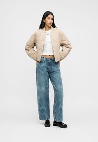 Μπεζ καπιτονέ μπουφάν με στρογγυλή λαιμόκοψη, λευκό κοντό τοπ και φαρδιά τζιν παντελόνια σε ανοιχτό μπλε με σκισμένο εφέ, ολοκληρωμένα με μαύρα παπούτσια.