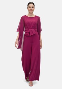 Fuchsia jumpsuit with flared trousers, a fitted waist tie, and flowing cape sleeves. Smooth fabric and minimalistic design.