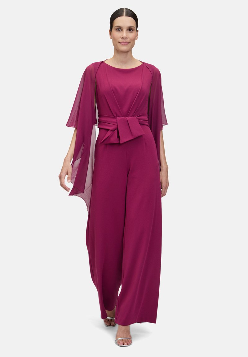 Fuchsia jumpsuit with flared trousers, a fitted waist tie, and flowing cape sleeves. Smooth fabric and minimalistic design.