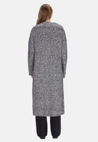 Persona con capelli lunghi che indossa un lungo cardigan grigio a maglia strutturata sopra pantaloni neri, in piedi di spalle contro uno sfondo semplice.
