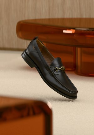 Čierny kožený mokasín s pozláteným kovovým detailom a značkou "HUGO", vystavený na béžovom koberci a v pozadí hnedý priehľadný nábytok.