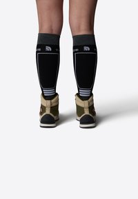 Medias negras hasta la rodilla con puños grises y rayas blancas, llevadas con botas de tobillo verdes y beis que presentan una suela texturizada y un detalle de logo.