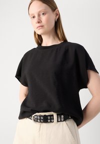 T-shirt noir à manches courtes avec une coupe relax, associé à une ceinture cloutée noire sur un pantalon crème. Texture de tissu lisse.