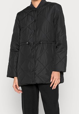 Veste matelassée noire avec boutons-pression et taille ajustable par cordon, portée sur un pantalon noir.