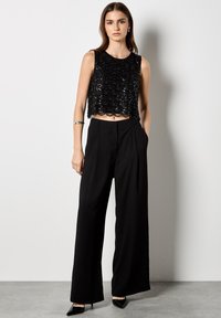 Top corto de lentejuelas negras con dobladillo en forma de escallón, combinado con pantalones de pierna ancha negros de cintura alta. La modelo lleva puestos unos tacones negros de punta.