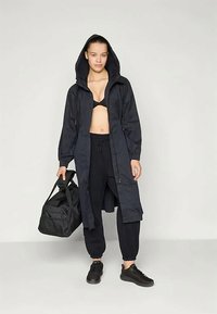 Lång svart huva jacka i lätt material, med frontdragkedja, tillsammans med svarta joggerbyxor och svarta sneakers.