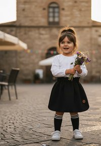 Dziecko w białej bluzce z długim rękawem i czarną spódnicą, trzymające dzikie kwiaty. Czarne skarpety do kolan w białe paski, białe sneakersy, tło z kostki brukowej.