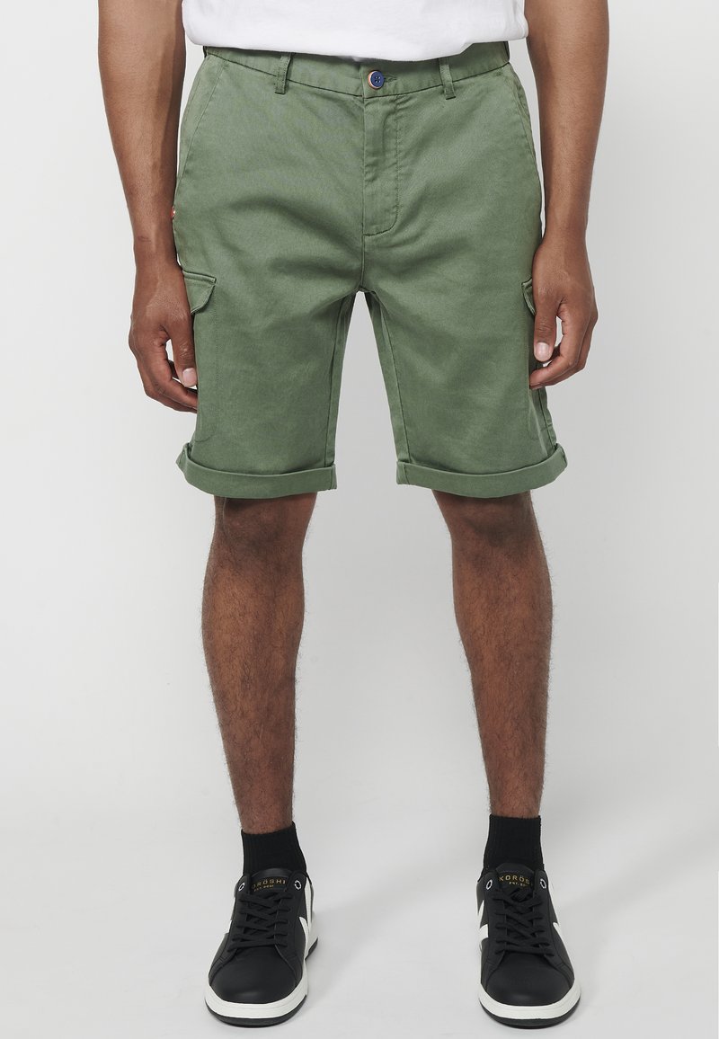 Grüne Baumwollshorts mit umgeschlagenen Säumen, seitlichen Taschen und einem Knopfverschluss. Kombiniert mit schwarzen Turnschuhen und knöchelhohen Socken.