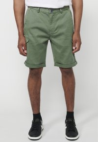 Man som har på sig olivgrön uppvikt cargoshorts, svarta strumpor och svarta sneakers som står mot en vit bakgrund.
