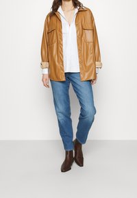 Hnědá koženková oversized košile s bundou a dvěma náprsními kapsami, v kombinaci s bílou košilí a modrými džíny, doplněná hnědými kotníkovými botami.