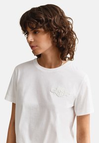 Jeune personne aux cheveux bouclés bruns, portant un t-shirt blanc avec un logo brodé discret sur la poitrine gauche, regardant sur le côté.