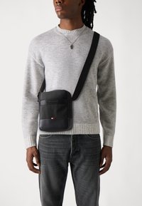 Black crossbody bag with textured surface, zip closure, and a small color block accent featuring red, white, and blue. Worn over a gray sweater.