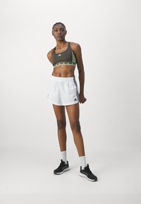 Sport bralette i mørkegrønn med grønne striper og logo, parret med hvite shorts med sidestriper, og svarte joggesko.