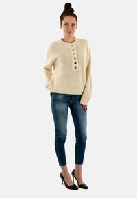 Maglione beige lavorato a maglia con scollo rotondo, cinque bottoni dorati e polsini a costine. Il modello indossa jeans skinny blu e tacchi alti neri.
