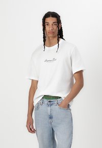 T-shirt bianca a girocollo con logo "Premium Frost" in nero, abbinata a jeans in denim azzurro e una cintura a quadri verdi. Maniche corte.