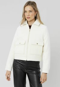 Veste blanche cropped en tissu doux, dotée d'un col, d'une fermeture éclair à l'avant et de deux grandes poches plaquées avec des boutons-pression.