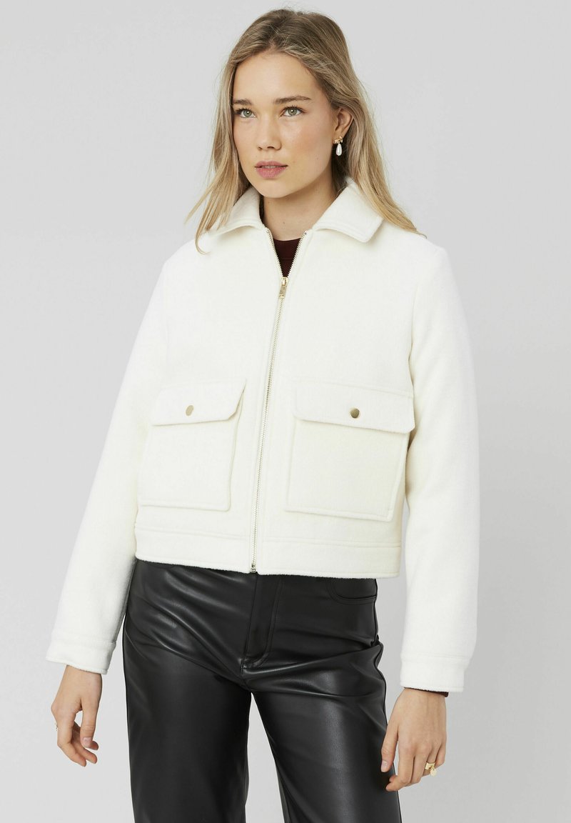 Veste blanche cropped en tissu doux, dotée d'un col, d'une fermeture éclair à l'avant et de deux grandes poches plaquées avec des boutons-pression.