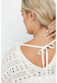 Vitt stickat linne med öppen ryggdesign och knytstängning, dekorerat med en strukturerad mönster, kompletterat av diskreta guldfärgade ringörhängen.