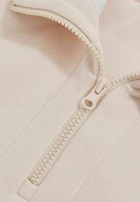 Tessuto beige chiaro con una cerniera liscia dotata di un'area di apertura rotonda, cuciture a contrasto e una finitura morbida e leggermente strutturata.