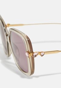 Gafas de sol de gran tamaño con un marco de plástico color marrón claro, lentes graduados en púrpura y varillas en tono dorado con acentos de cristal rosa.