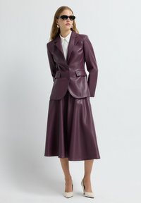 Kvinde iført sorte solbriller, hvid bluse, plumfarvet læderblazer og matchende midi-nederdel, stående med hænderne bag ryggen, beige hæle.