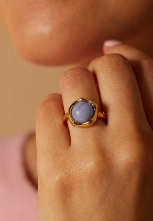 Hand met gouden ring met een ronde, gepolijste blauwe steen, gehouden bij het lippengebied met een lichte huid en een neutrale achtergrond.