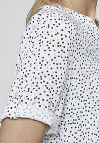 Weißes Hemd mit einem Tupfenmuster aus schwarzen und hellblauen Punkten, hochgekrempelte Ärmel und eine Knopfverzierung nahe der Manschette.