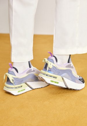 Feet in white pants and socks wearing purple, yellow, and white chunky sneakers with visible air cushions, standing on a mustard-colored floor.