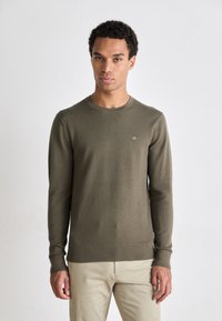 Maglione a maglia texturizzata verde oliva, con maniche lunghe e collo rotondo; presenta un piccolo logo bianco sul petto; abbinato a pantaloni beige chiaro.