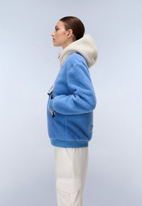 Blue fleece jacket with a cream hood, featuring front pockets and a blue trim at the hem. Smooth texture with a relaxed fit.