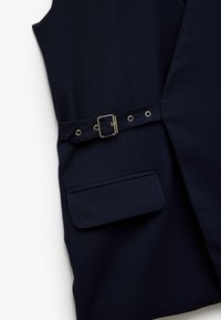 Marineblaues ärmelloses Kleidungsstück mit strukturiertem Design, ausgestattet mit einem Gürtel mit rechteckiger Schnalle und einer einzigen Fronttasche. Glatte Stoffoberfläche.