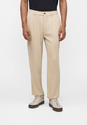 Hombre con pantalones beige plisados y zapatillas beige con suelas negras, de pie sobre un fondo blanco liso.