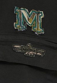 Svart keps med en broderad "M"-logotyp i grönt och beige med fransar, samt en örn-design med texten "Air Force" i kontrasterande broderi.