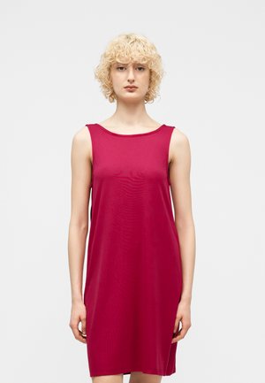 Kõrgetel blondidel lokkis juustel naine, kes kannab varrukateta, põlveni ulatuvat punast kleiti ja seisab ühetaustase helehall tausta ees.