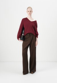 Burgunderfarbener, übergroßer Pullover mit überschnittenen Schultern, kombiniert mit hoch taillierten, braunen, gefalteten Hosen und einer schwarzen Handtasche aus Lackleder.