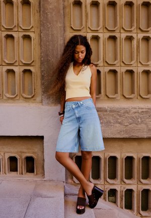 Jeune femme aux longs cheveux bouclés portant un haut beige sans manches, un short en jean et des sandales compensées noires, appuyée contre un mur texturé.
