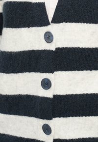 Detailný záber na pletenú cardigan s čierno-bielymi horizontálnymi pruhmi, s tromi viditeľnými tmavými gombíkmi pozdĺž prednej strany.